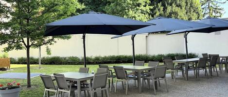 An outdoor patio with tables and chairs, covered by umbrellas, and surrounded by greenery.