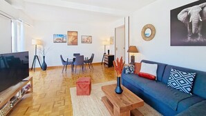 A modern living room with a flat-screen TV, a blue sofa, a coffee table, and a dining area with chairs and a table. - N&J - TROPI AZUR - Terrace - Place du Pin  (Niza)