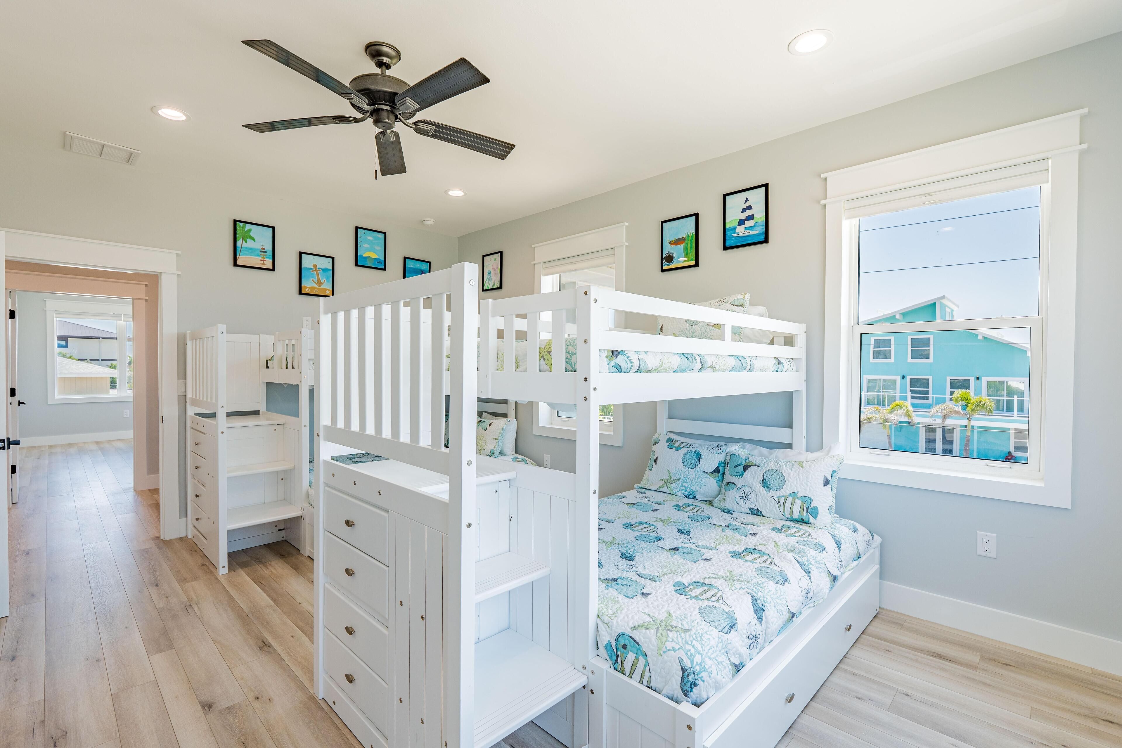 A child’s bedroom with bunk beds, a ceiling fan, and a window with a view of a blue house.