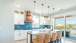 A modern kitchen with white cabinets, a blue backsplash, and a large island with wicker chairs. - Family Beach House (Fort Myers Beach)