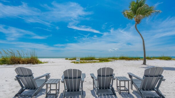 A beach with four Adirondack chairs and a table, a palm tree, and a clear blue sky. - Family Beach House (Fort Myers Beach)