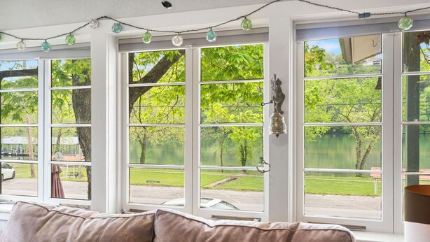 A room with large windows, a brown leather couch, and a view of a green landscape with trees and a body of water. - The Raven's Roost Lake View Retreat 3bd 2ba (Rockaway Beach)