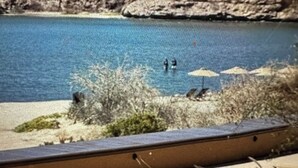 Blick durch ein Fenster auf einen Poolbereich mit klarem, blauem Pool, Sandstrand und etwas Grün.
