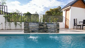 Ein Pool mit einem Wasserfall aus Stein, umgeben von einem schwarzen Zaun und einem weißen Gebäude mit Holzdach.