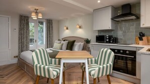 Un dormitorio compacto con cama, mesita de noche y ventana con cortinas. Junto a él, una kitchenette con microondas, tostadora y horno. Hay sillas a rayas y una pequeña mesa en la habitación.