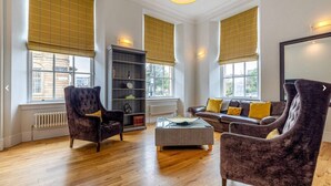 A spacious living room with a dark velvet sofa, a matching armchair, a bookshelf, and large windows with blinds.