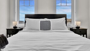 Una cama bien hecha con sábanas blancas y un cojín estampado, flanqueada por dos mesitas de noche con lámparas, en una habitación con ventanas amplias que ofrecen vistas del cielo y edificios.