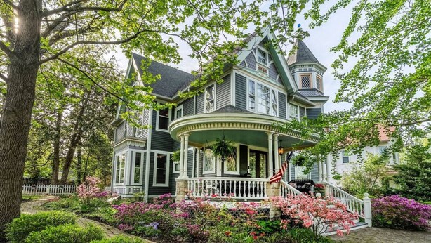 Une grande maison à deux étages, dotée d’une véranda qui fait le tour de la maison, et entourée d’un jardin bien entretenu où poussent différentes espèces de plantes à fleurs.