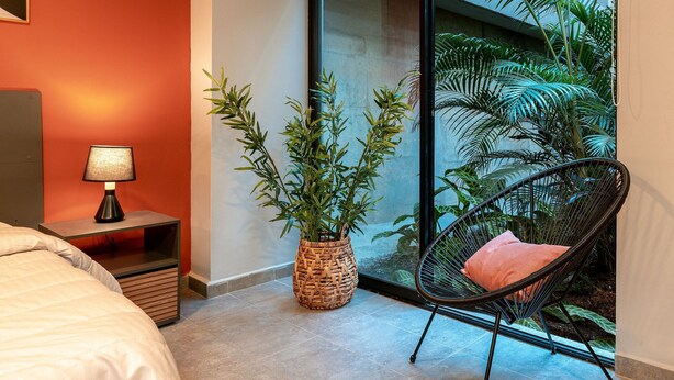 A hotel room with a bed, a nightstand with a lamp, a wicker plant, a black chair with a cushion, and a view of greenery through a glass door.