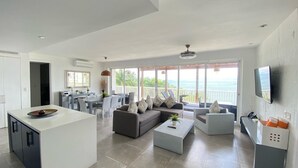 A spacious living area with a kitchen, dining table, and seating arrangements. A large window offers a view of the ocean. - Apartamento Vista Mare 9.102 (Samana)