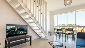 A modern living room with a TV on a black stand, a staircase, a dining area with chairs and a table, and large windows offering a view. - Hyères - Apartment T5 - Sea and salt water view - Refurbished (Hyères)