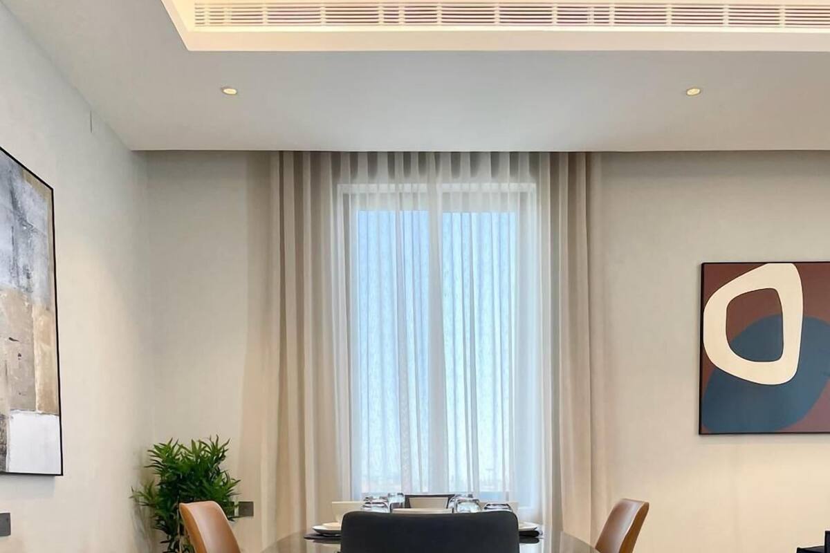 A modern dining area with a glass table, two brown chairs, and a large window with sheer curtains.