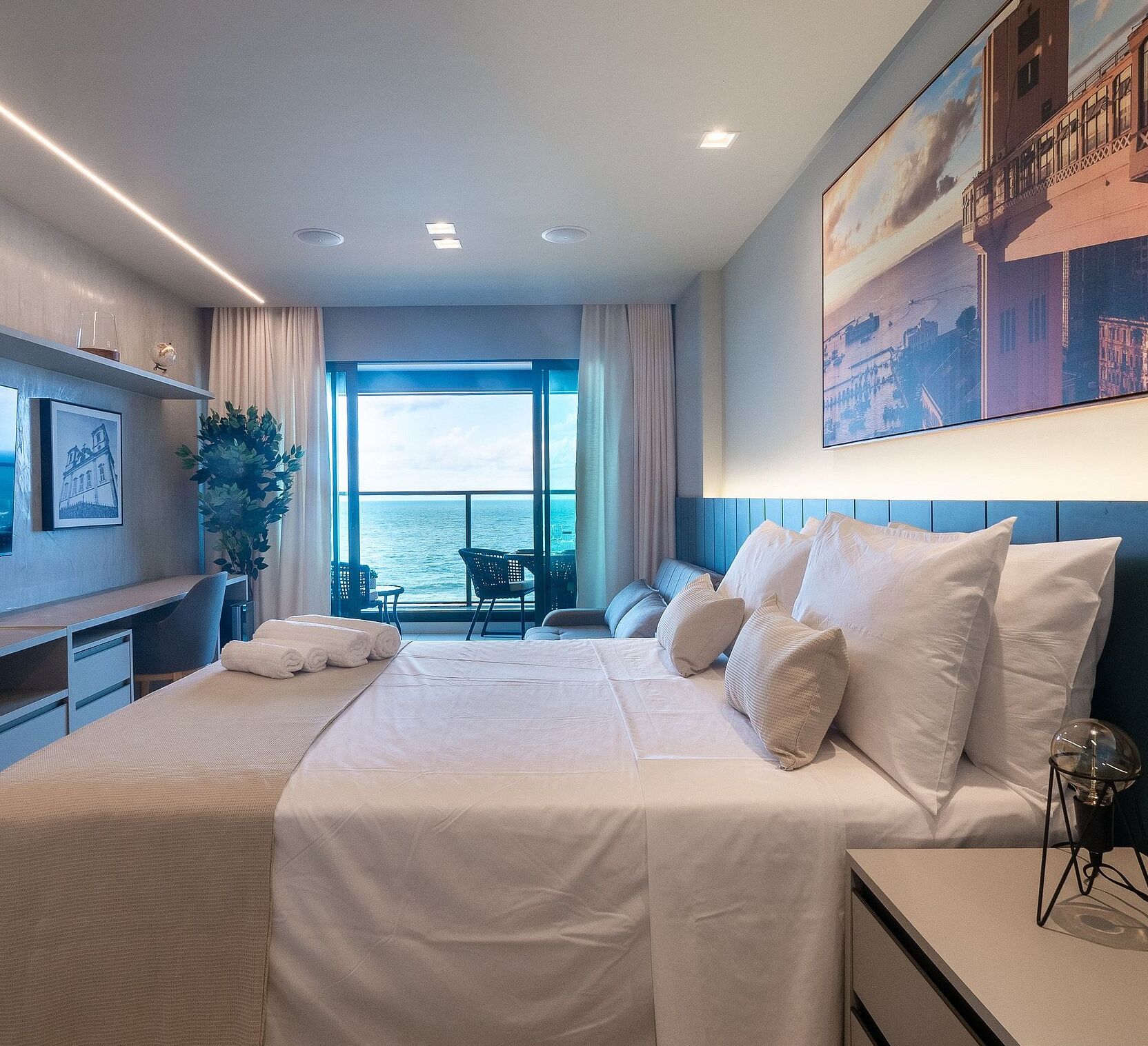 A modern hotel room with a large bed, a desk, and a view of the sea.