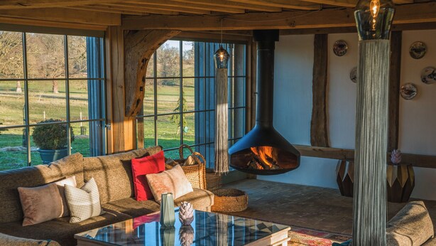 A cozy living room with a large window, a fireplace, and a high wooden ceiling. - Wilderness Reserve - Cartshed (Halesworth)
