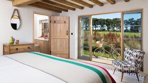 A bedroom with a large bed, wooden ceiling beams, a sliding glass door leading to a garden, and a chair by the window. - Wilderness Reserve - Cartshed (Halesworth)