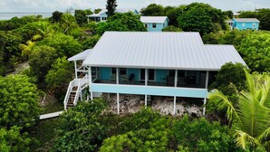 A coastal house with a white roof and light blue exterior, surrounded by lush greenery and a clear view of the sea.