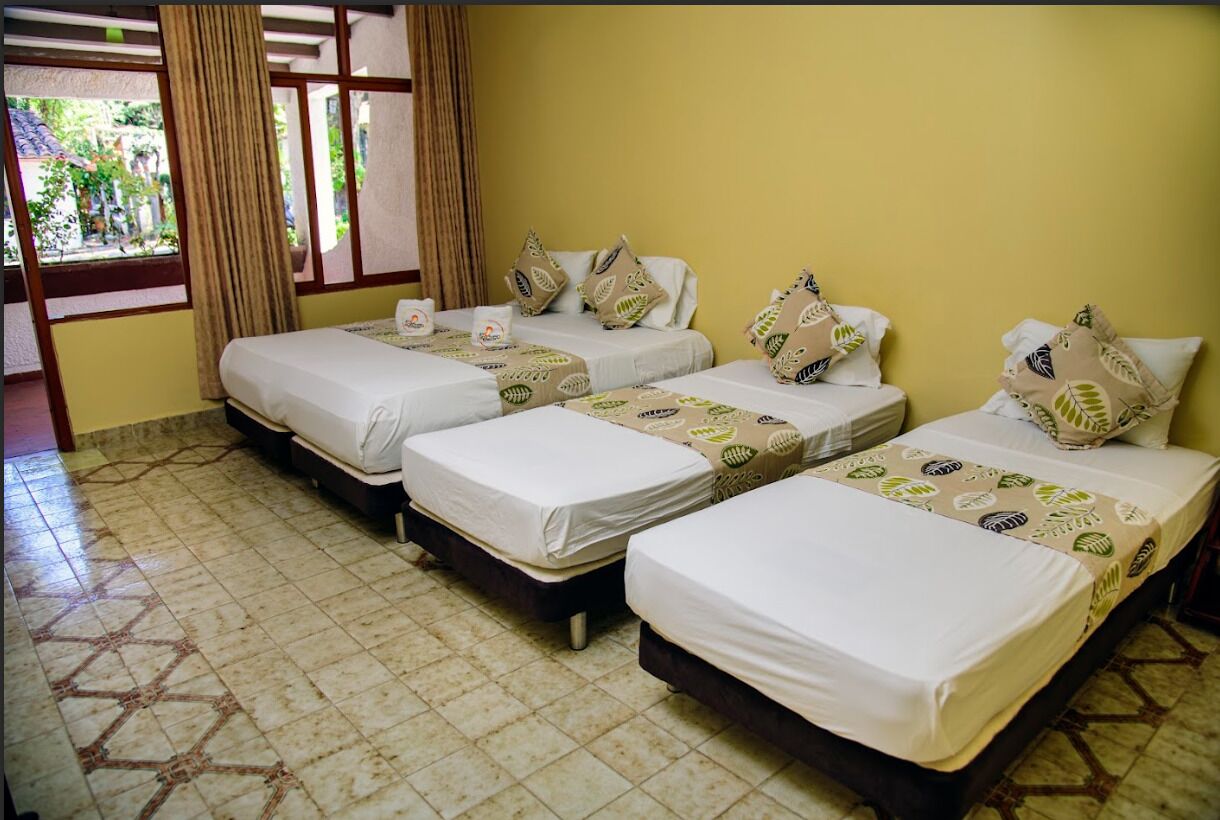 Habitación de hotel con tres camas individuales, cada una con una colcha estampada y almohadas a juego. El piso es de baldosas y hay una ventana con cortinas.