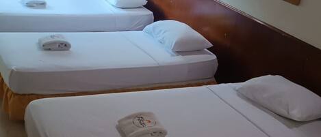 A hotel room with three single beds, each with a neatly made white bedspread and a towel with a logo. The room has a wooden headboard, a window with curtains, and a framed picture on the wall.