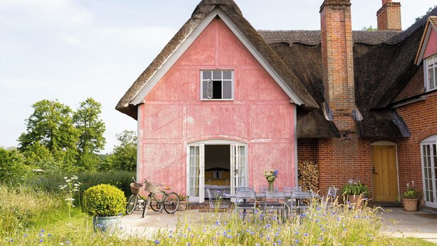Une maison rose avec un toit de chaume, une porte en bois et un petit porche avec une table et des chaises.