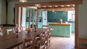Un coin repas rustique avec des chaises et une table en bois, une cuisine aux tons turquoise dotée d’un grand îlot central, et des poutres en bois apparentes au plafond.