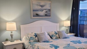 Ein Schlafzimmer mit einem Bett, Nachttischen, einem Deckenventilator, einem Gemälde und Blick auf Gebäude durch ein Fenster.