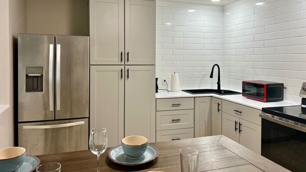 A modern kitchen with white cabinets, a stainless steel refrigerator, a black microwave, and a wooden dining table with blue plates and glasses. - Spacious 1-bedroom guesthouse in lovely Wiggins with WiFi, AC (Wiggins)