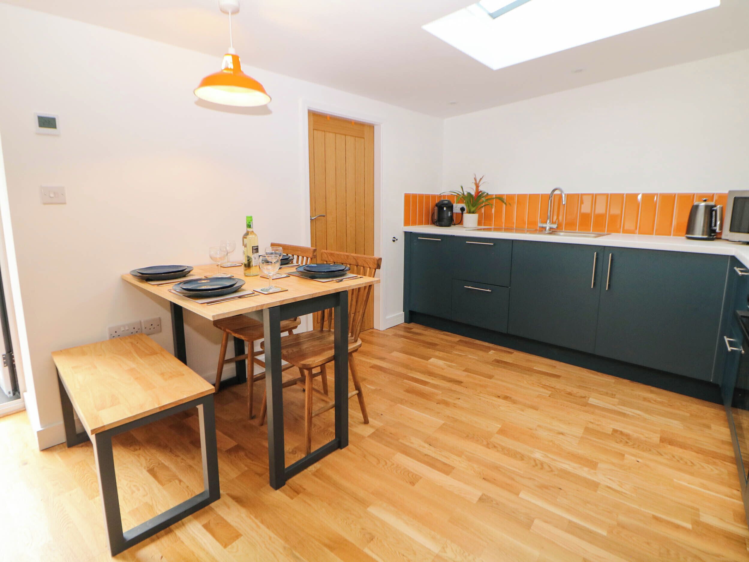 A compact kitchen with wooden flooring, a dining table with chairs, a bench, and a skylight above.