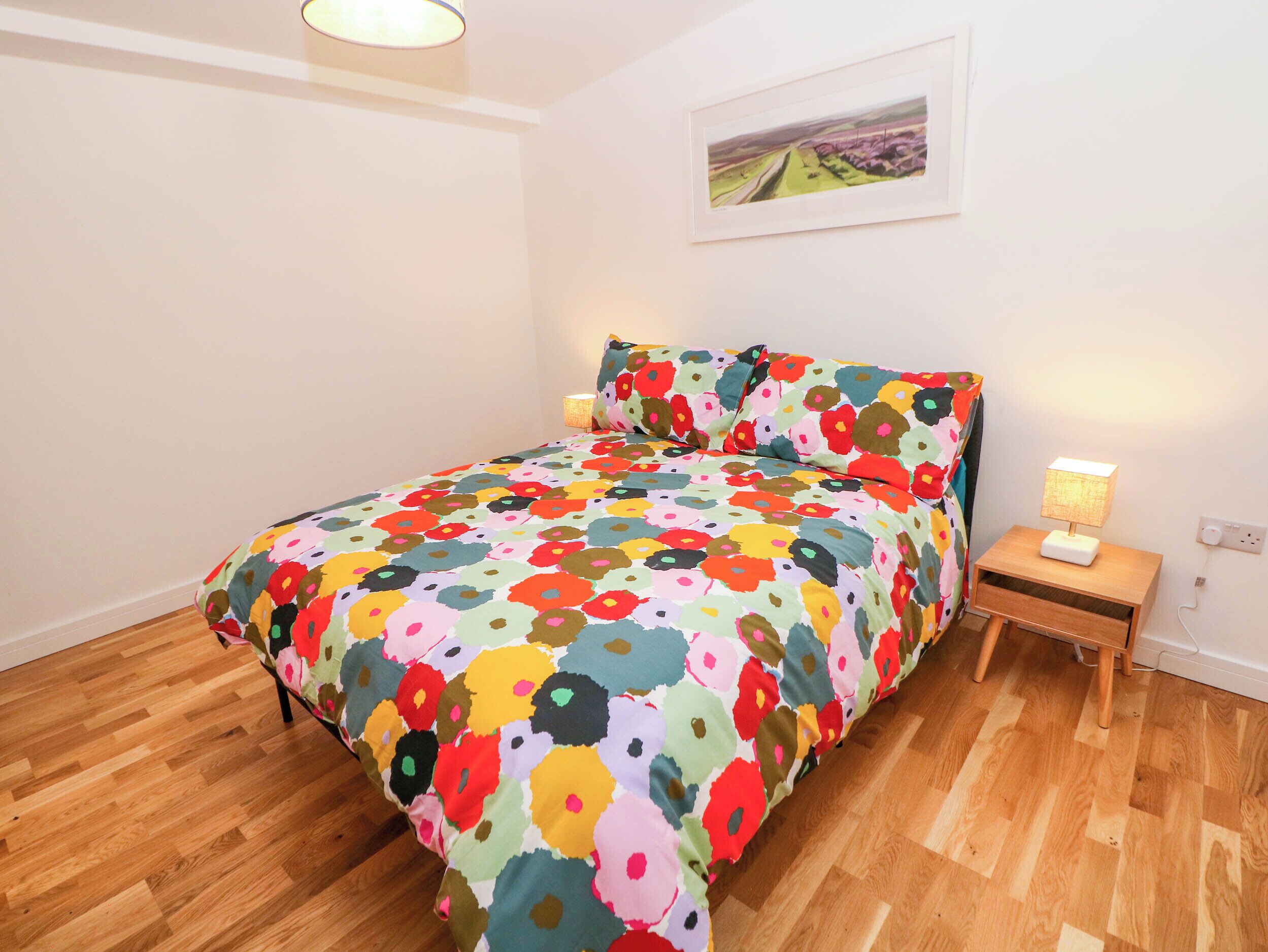 Un dormitori amb llençols estampats florals, terres de fusta, una tauleta de nit amb una làmpada i un quadre amb marc a la paret.