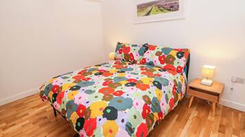 Un dormitori amb llençols estampats florals, terres de fusta, una tauleta de nit amb una làmpada i un quadre amb marc a la paret.