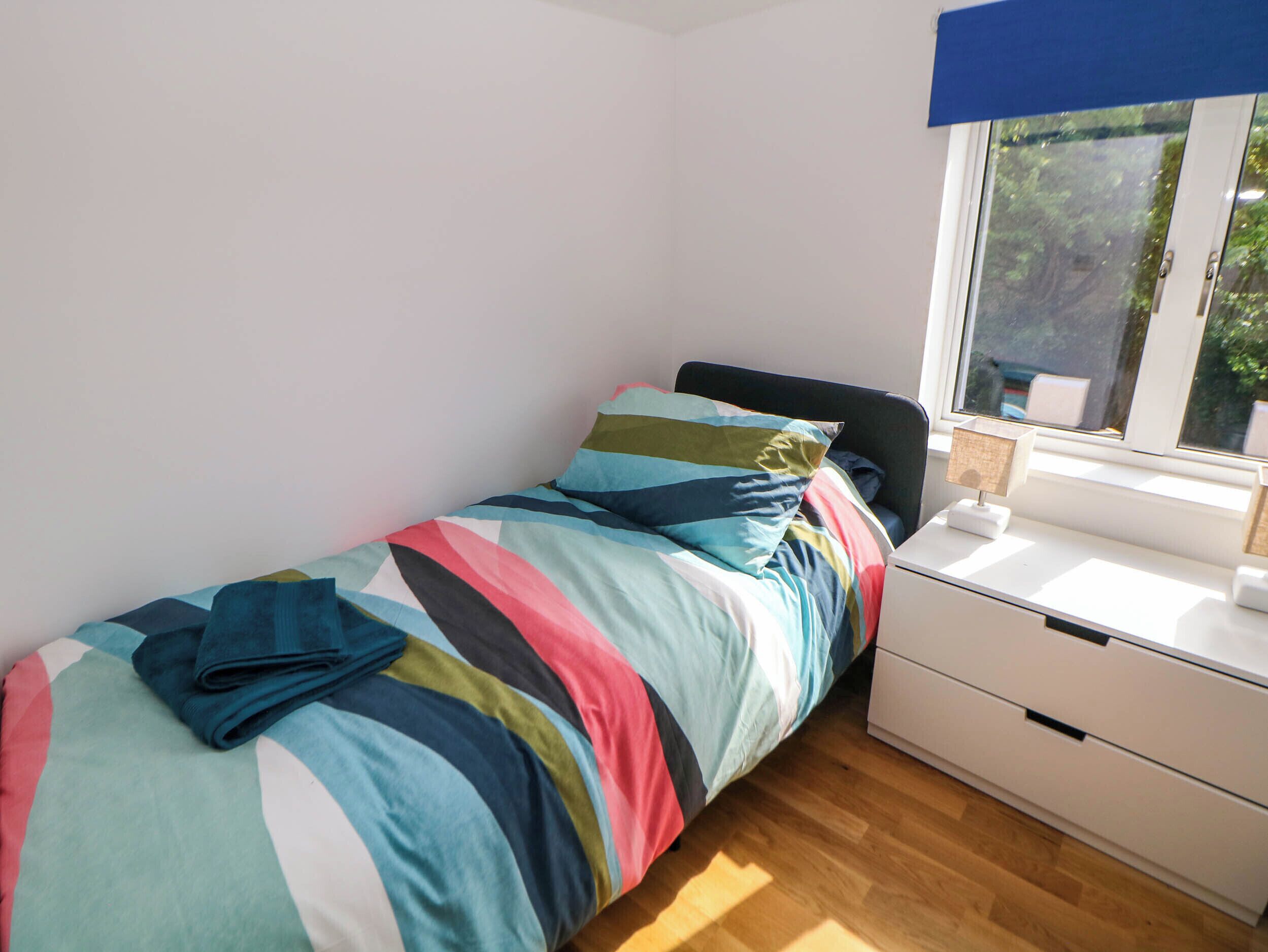 A small, neatly made bed with colorful bedding, a bedside table with a lamp, and a window with a view of greenery.
