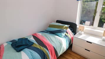 A small, neatly made bed with colorful bedding, a bedside table with a lamp, and a window with a view of greenery.