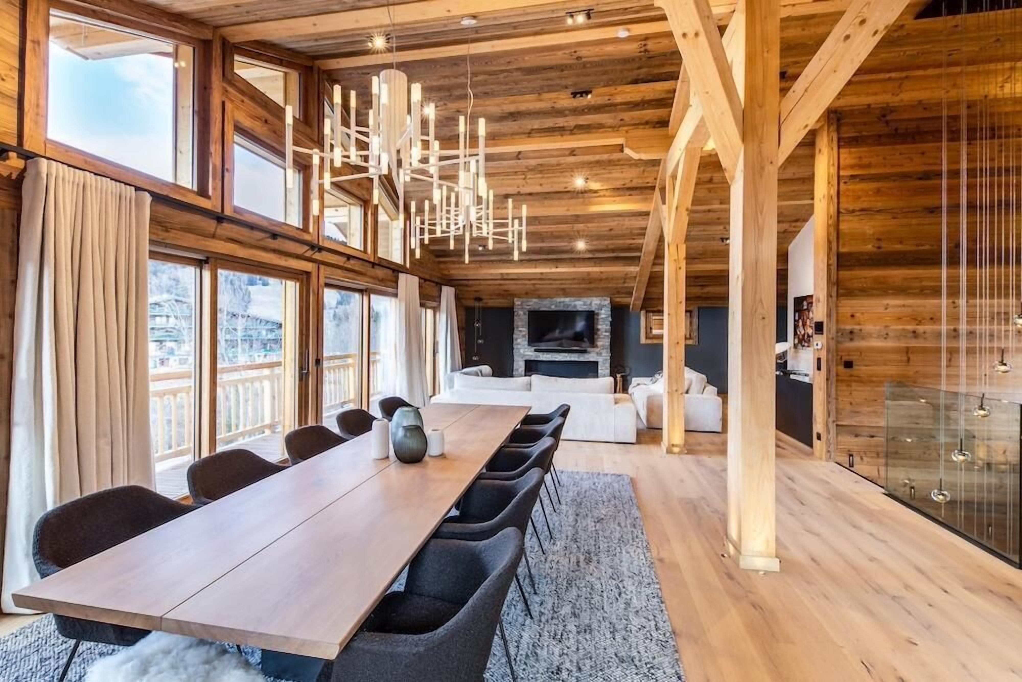 Una habitación amplia con una larga mesa de madera, asientos cómodos, una chimenea y ventanas grandes que ofrecen vistas al exterior.