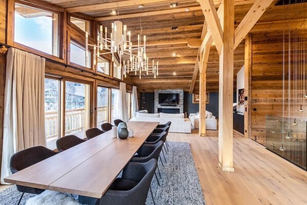 A spacious room with a long wooden table, comfortable seating, a fireplace, and large windows offering a view of the outdoors.