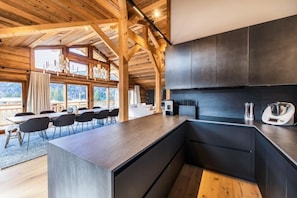 Una cocina moderna con armarios oscuros y una isla de madera, ventanas amplias y una zona de comedor con sillas y mesa.