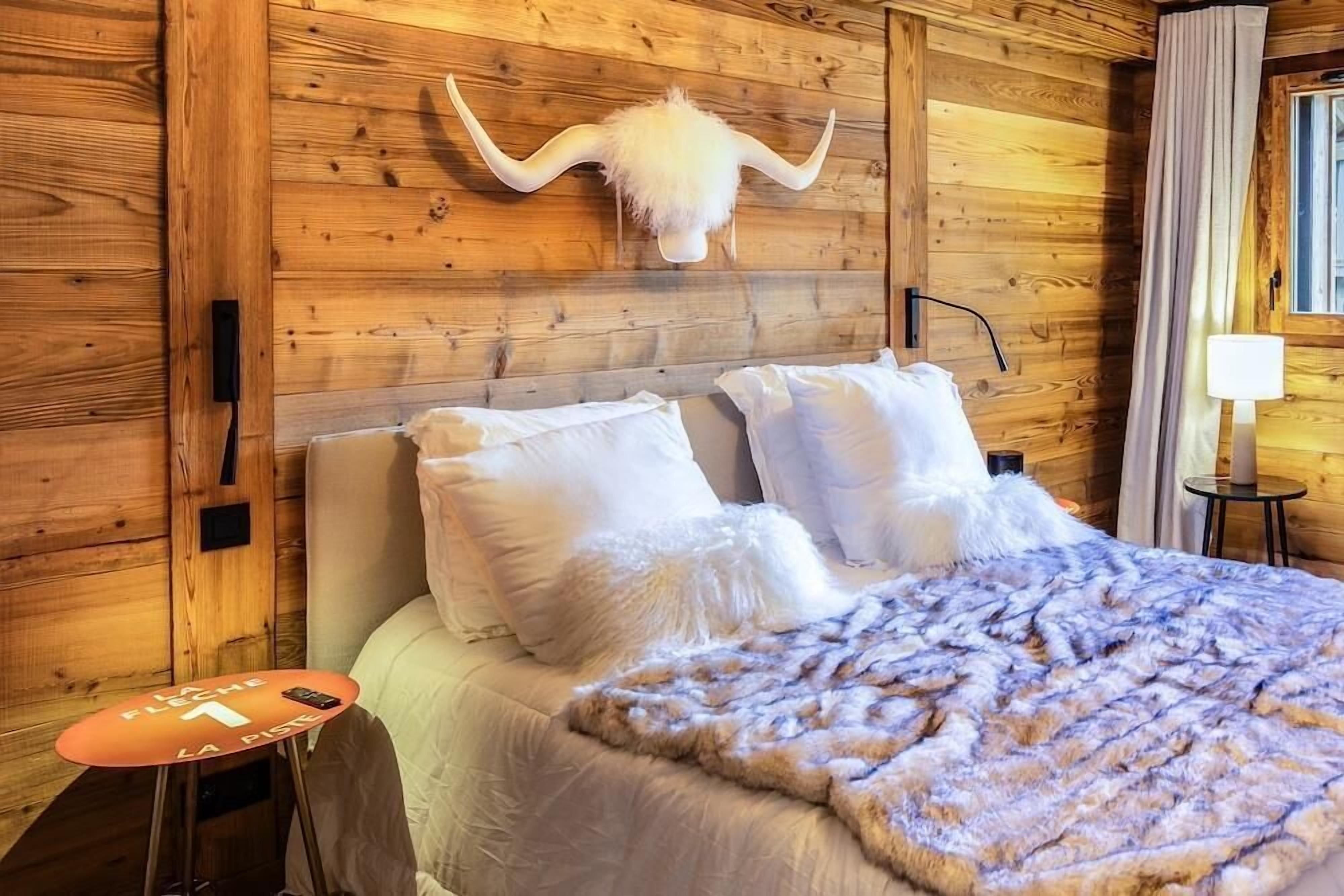 A wooden-paneled room with a bed, white pillows, a fluffy blanket, and a mounted animal head on the wall.