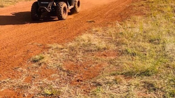 Deux personnes conduisent des quads sur un chemin de terre, avec des arbres et un ciel dégagé en arrière-plan.