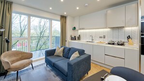 A modern kitchen with a herringbone backsplash, a dining area with a sofa and chairs, and a large window with a view of trees. - Stylish Apartment on Colindale Avenue (London)