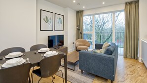 Een moderne woonkamer met een eethoek, een bank en een televisie. Er hangen ingelijste foto’s aan de muur, een raam met gordijnen, en een balkon met een hekwerk.