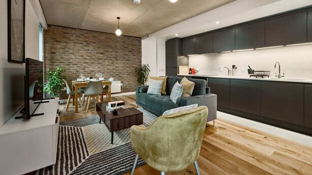 Una sala de estar moderna con comedor, un sofá gris, un sillón verde y una cocina con armarios oscuros y encimera blanca.