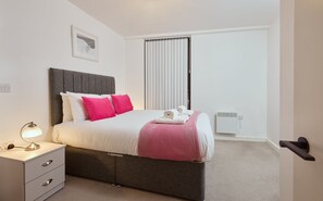 Un dormitorio moderno con un cabecero tapizado gris, ropa de cama blanca y un plaid rosa. Hay una mesita de noche con una lámpara, un equipo de aire acondicionado blanco y una puerta con pomo negro.