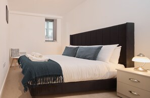 Una cama bien hecha con un cabecero oscuro, una lámpara de noche y una silla con una manta.
