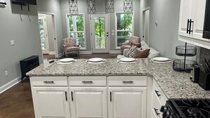 Une cuisine moderne avec des armoires blanches, des comptoirs en granit et une cuisinière à gaz. Des luminaires suspendus sont placés au-dessus de l’îlot central, et une grande fenêtre apporte une lumière naturelle.