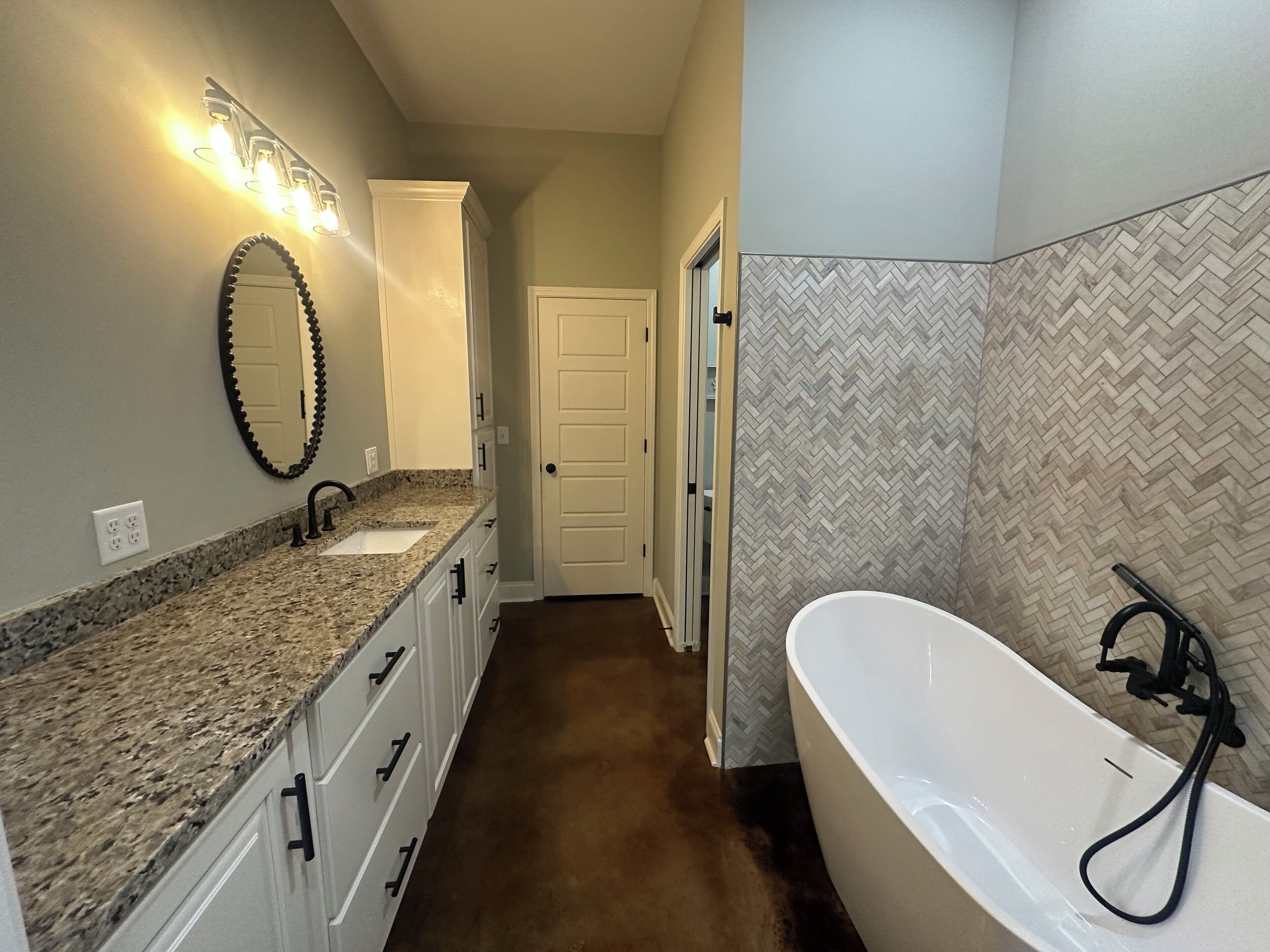 A bathroom with a granite countertop, a freestanding bathtub, a mirror with a beaded frame, and a herringbone-patterned tile backsplash.