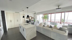A modern kitchen with a central island, a dining area with a white sofa, and a balcony with a view of palm trees. - APARTAMENTO VISTA MARE 9-202 (Samana)