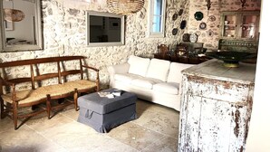 Une pièce rustique aux murs de pierre, avec un banc en bois, un canapé blanc et un coin repas comprenant une table et des chaises en bois.