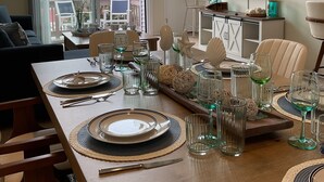 A dining area with a long table set for four, a television mounted on the wall, and a view of the outside through large windows. - Newly Furnished Sun Drenched Duplex Private Deck Steps to the Beach & Boardwalk! (Seaside Heights)