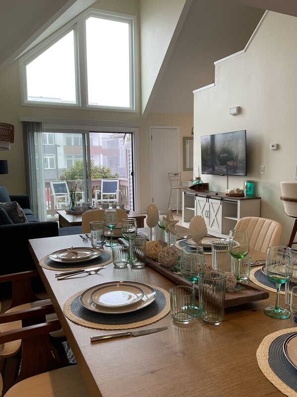 A dining area with a long table set for four, a television mounted on the wall, and a view of the outside through large windows.
