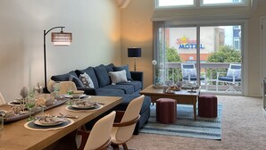 A spacious living area with a dining table set for four, a ceiling fan, and a view of a motel sign through a large window. - Newly Furnished Sun Drenched Duplex Private Deck Steps to the Beach & Boardwalk! (Seaside Heights)