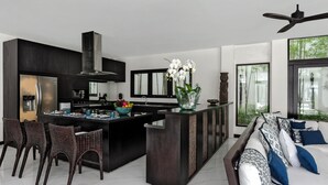 A modern kitchen with a central island, dark cabinetry, and a dining area with wicker chairs. - Luxevacay Anaya Manis Villa lvid3 (Badung)