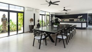A modern dining area with a long table, black chairs, ceiling fans, and a view of greenery through large windows. - Luxevacay Anaya Manis Villa lvid3 (Badung)